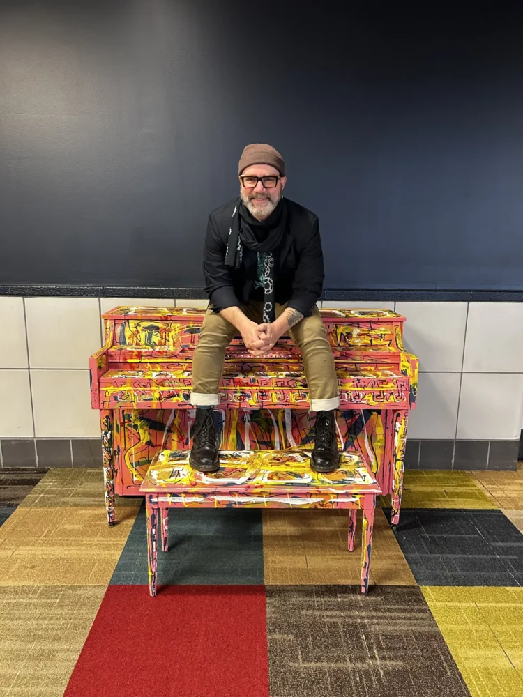Artist seated on a brightly painted abstract piano in the Pittsburgh International Airport.